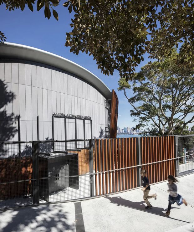 Exterior view of a contemporary curved-roof civic or educational building, clad in pale vertical panels with a prominent timber-toned slatted fence and dark metal security mesh. A sign is mounted above the entrance, while mature trees frame the building and a distant urban skyline is visible beyond. Two people are walking along the paved foreground.