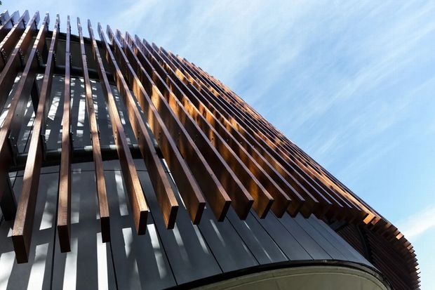 Low-angle architectural exterior view of a contemporary multi-storey building facade. Dense vertical timber-toned fins project outward over dark glazing and smooth grey cladding, creating strong linear shadows and a dramatic rhythm against a clear blue sky. The image focuses on facade articulation and external envelope rather than an interior space.