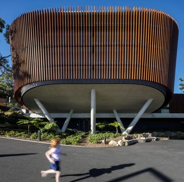A striking contemporary multi-storey building viewed from the front, with a rounded projecting upper volume wrapped in closely spaced vertical timber-toned fins. The upper level is supported by angled structural columns and a central round column over a broad curved concrete soffit. Dark glazing and horizontal floor divisions are visible behind the screen, with landscaped planting and a paved driveway in the foreground.