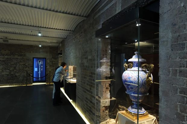 Dimly lit museum or gallery interior housed within a rugged masonry space. Dark brick and stone walls frame illuminated glass display cases containing ornate decorative objects, including a large blue-and-white urn. A curved ribbed metal ceiling and concealed linear lighting create a dramatic exhibition atmosphere, with a visitor viewing a display counter in the background.
