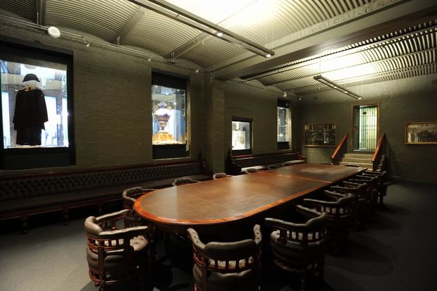 A dark, refined commercial or institutional meeting room with a large polished wooden conference table surrounded by ornate upholstered chairs. The space features exposed dark masonry walls, black-framed windows, built-in banquette seating, framed artwork, and a ribbed industrial-style metal ceiling with linear lighting. Display windows and a raised area with steps are visible along the rear wall.