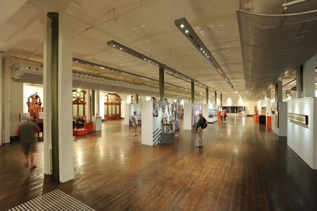 A spacious museum or gallery interior with polished dark timber flooring, tall pale columns, exposed services and ductwork, suspended linear track lighting, and open exhibition areas with display cases and artifacts. Visitors move through the bright, expansive space. The architecture combines an industrial ceiling and structural system with a refined heritage-like gallery fit-out.