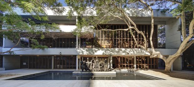 A contemporary multi-storey building facade arranged around a rectangular reflecting or swimming pool. The white and grey rendered structure has extensive dark timber-framed glazing, deep covered balconies, vertical screening, and a mature tree integrated into the forecourt. A sculptural feature wall or artwork sits beside the pool, creating a shaded courtyard-like setting.