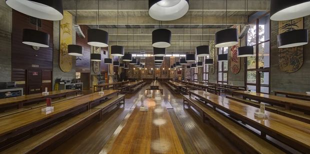 A large industrial-style communal dining hall or food-hall interior with long polished timber tables and benches arranged in rows. The space features a high exposed concrete ceiling, extensive dark timber wall panelling, large windows, numerous suspended black-and-white pendant lights, and decorative wall artworks. The symmetrical perspective emphasizes the length and generous scale of the room.