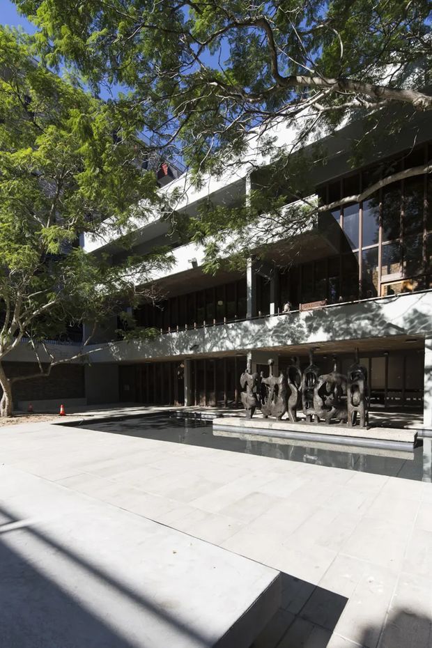 Exterior view of a multi-storey modern institutional or commercial building set among mature trees. The facade combines broad white concrete planes, dark vertical window framing, recessed glazing and projecting horizontal levels. A paved forecourt with a shallow rectangular reflecting pool and a row of sculptural figures occupies the foreground.
