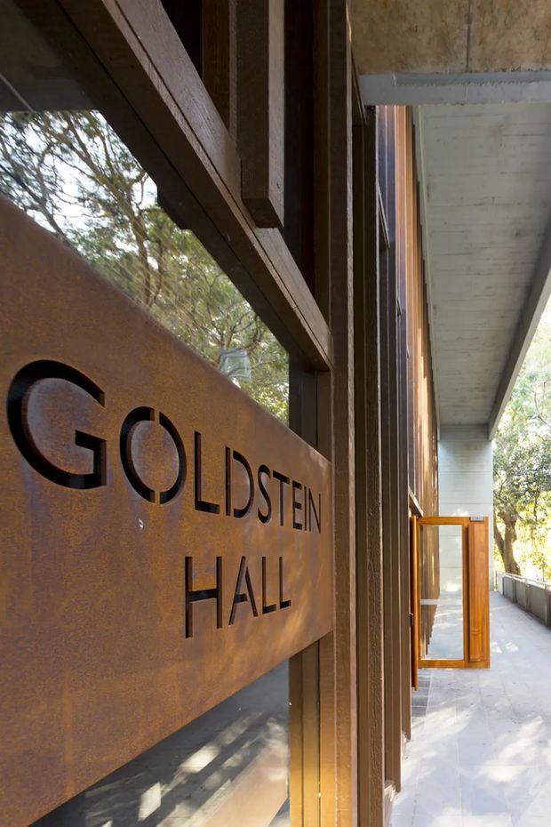 Close-up architectural view of a building entrance or corridor featuring a large rust-toned metal sign reading “GOLDSTEIN HALL.” Dark timber framing, exposed concrete soffit, timber-framed glazing, and a long open walkway are visible, with trees beyond the building. The image emphasizes the exterior architectural detailing and signage.
