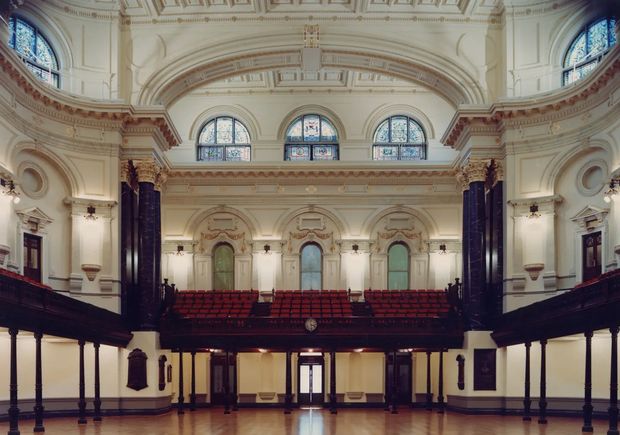 A grand historic interior hall with symmetrical balconies, ornate classical plasterwork, decorative cornices, arched detailing, stained-glass windows, dark polished columns, and rows of red auditorium seating. The polished timber floor and formal monumental composition suggest a heritage theatre, concert hall, or civic assembly space.