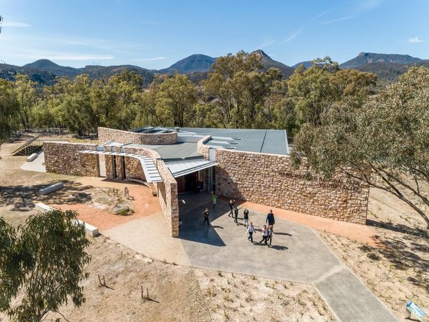 Aerial view of a low-profile contemporary building set within a dry, wooded mountain landscape. The structure has extensive rough natural stone masonry walls, a broad flat roof, curved and angular roof forms, and a paved entrance forecourt with several people for scale.
