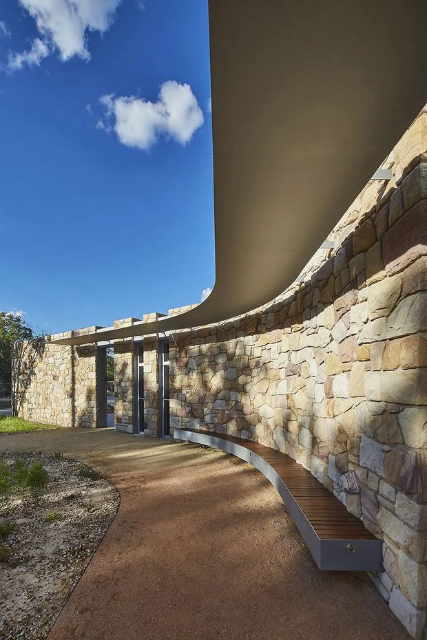 Exterior architectural view of a curving stone-clad wall beneath a dramatic sweeping roof or canopy. A curved timber bench follows the wall, while a paved walkway leads through a sheltered open-air passage. The composition highlights organic curves, substantial masonry, dark structural supports, and a strong contrast between warm stone and blue sky.