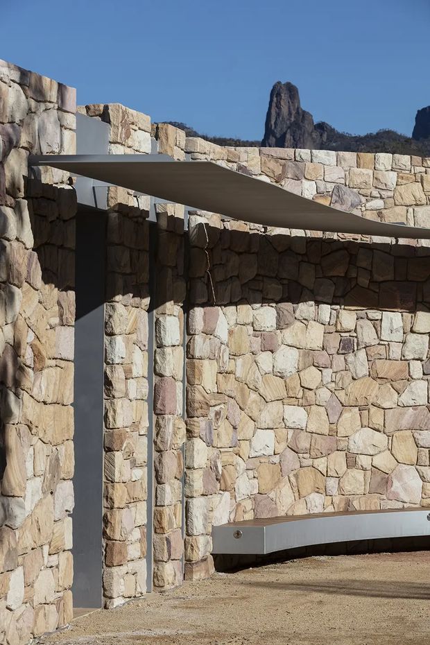 Exterior architectural detail featuring robust irregular natural-stone masonry walls and slender dark metal structural elements. A broad curved metal canopy projects across the courtyard-like space, with a low metal bench or ledge along the stone wall. Distinctive rugged rock formations are visible beyond the building, under a clear blue sky. The composition emphasizes a contemporary structure integrated into a rocky landscape.