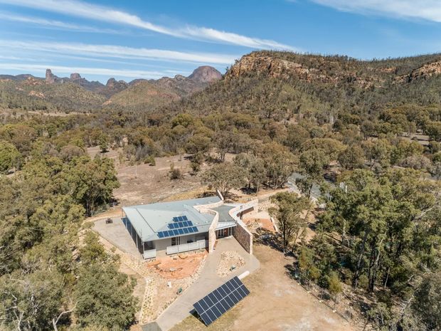 Aerial view of a low-profile modern building set within a dry, wooded landscape and rugged rocky mountain range. The building has a pale metal roof with rooftop solar panels, additional ground-mounted solar panels, angular roof forms, stone-clad walls, and surrounding paved paths and terraces. The scene is sunny and remote, with expansive bushland and dramatic rock formations.