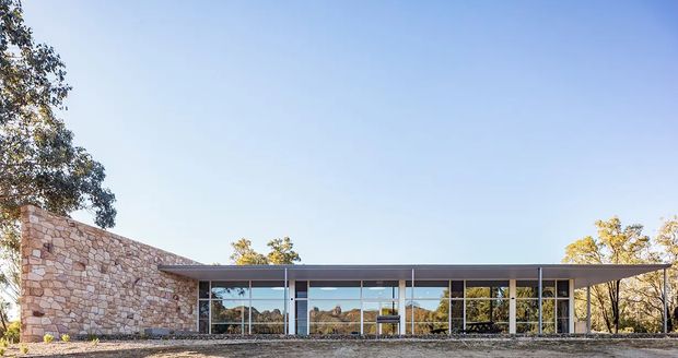 Low, single-storey modern pavilion with extensive floor-to-ceiling glazing, a broad flat roof and a substantial warm-toned stone masonry wall. The building sits in a natural, lightly wooded setting and is photographed from outside under a clear sky.