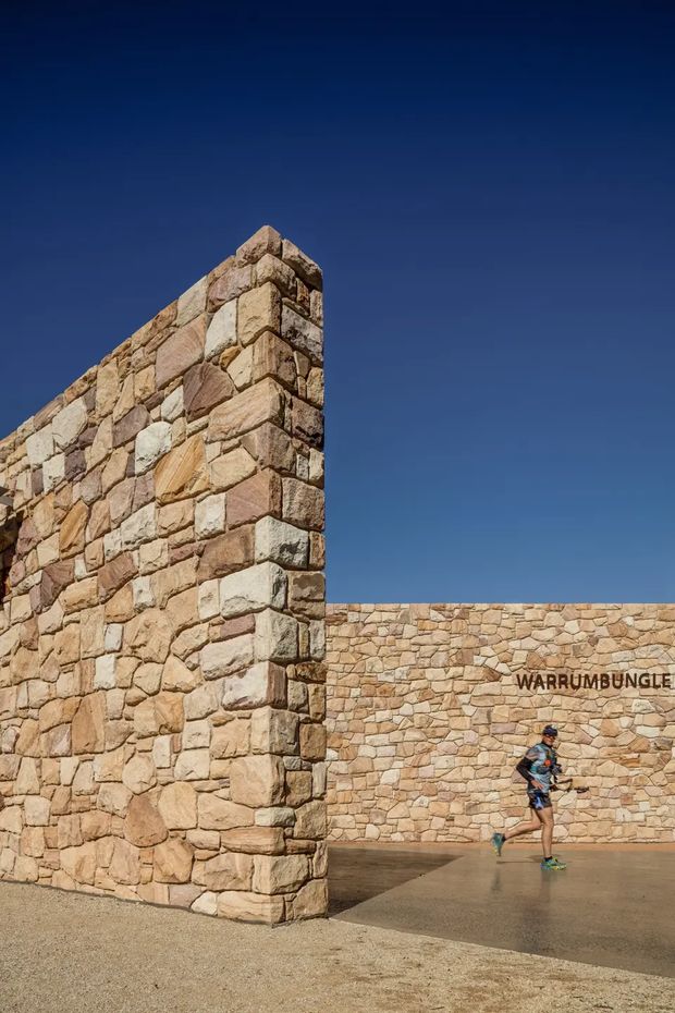 Exterior architectural photograph of a monumental dry-stone or masonry wall installation forming a tall projecting corner and low boundary wall. The walls use irregular warm-toned sandstone blocks in beige, tan, pink and brown, set against a deep blue sky. A paved forecourt and a small runner provide scale. The word “WARRIUMBUNGLE” is mounted on the rear wall.