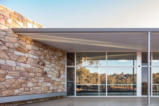 Exterior architectural photograph of a low, modern pavilion-like building with a broad flat roof and extensive floor-to-ceiling glazing. The facade combines irregular natural stone walling with slim light-coloured metal-framed sliding glass doors and windows. A covered terrace or entry zone occupies the foreground, with a slender support post and reflections of trees and rocky terrain in the glazing.