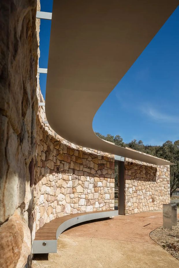 Exterior architectural view of a sculptural curved canopy and integrated bench wrapping around a rough stone masonry wall. The design uses warm irregular natural stone, smooth light-coloured curved soffits and metal supports, with a paved forecourt and open blue sky visible beyond. The composition highlights the canopy's sweeping form and the relationship between contemporary structure and rugged masonry.