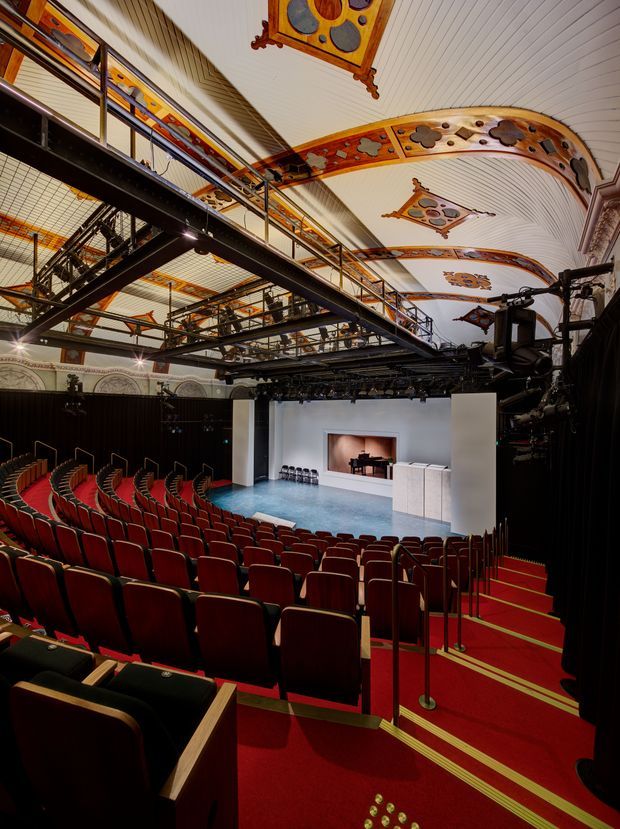 Interior view of an ornate theatre or performance auditorium, looking down over rows of red upholstered seats toward a small stage. The space features elaborate painted and carved ceiling panels with curved decorative beams, a dark technical lighting grid and catwalks overhead, patterned wall detailing, red carpeted aisles, and accessibility handrails. The stage includes a piano and simple white podium or set pieces.