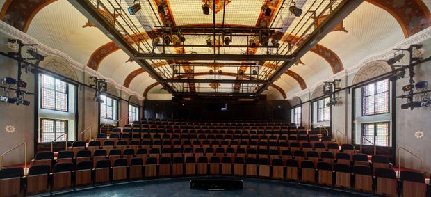 A grand historic auditorium viewed from the stage, with curved rows of red upholstered theatre seats, ornate decorative wall and ceiling detailing, tall leaded-style windows, and a large suspended lighting and rigging grid overhead. The space has a formal, richly ornamented character and appears designed for performances or lectures.