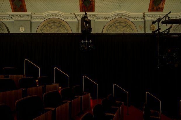 Dark interior view of a historic theatre or auditorium, showing tiered rows of upholstered seats with timber-toned side panels, red carpeted aisles, slim brass-coloured handrails, and a black stage curtain. Ornate plaster cornices, decorative arched panels, stained-glass-like ceiling details, and theatrical lighting equipment are visible above and around the auditorium.