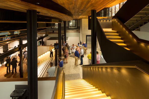 Interior view of a multi-level public or commercial building, looking across an open atrium with visitors gathered below. Dramatic illuminated staircases with dark metal sides and warm timber treads connect the levels. The space features exposed timber ceiling and soffit surfaces, black structural columns and balustrades, large areas of glazing, and a terrazzo or speckled floor. The overall design is contemporary, highly layered, and warmly lit.