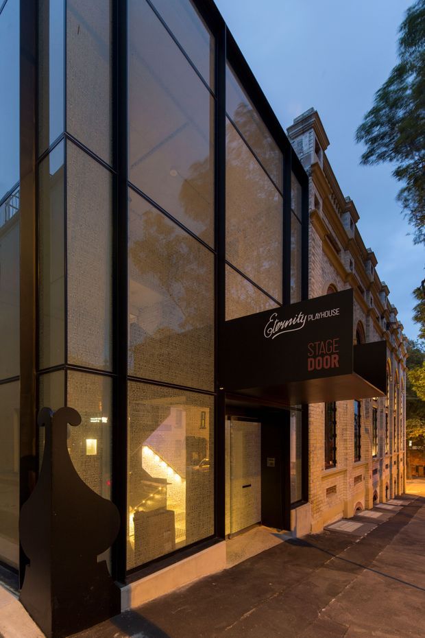 Dusk exterior view of a contemporary theatre or performance venue entrance beside a retained historic masonry building. The modern facade uses tall dark-framed glazed panels with translucent patterned screening, a projecting black canopy with the signage “Eternity Playhouse” and “Stage Door,” and a sculptural silhouette sign. Warm interior lighting is visible through the glazing, while the adjacent heritage-style facade features ornate cornices, pilasters, and arched openings.
