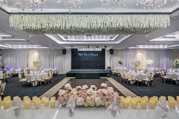 A grand formal event hall styled for a wedding or gala, with round banquet tables, white and gold seating, layered drapery, floral centerpieces, a raised stage, dramatic floral ceiling installation, crystal chandeliers, and candlelit floral decor in the foreground.