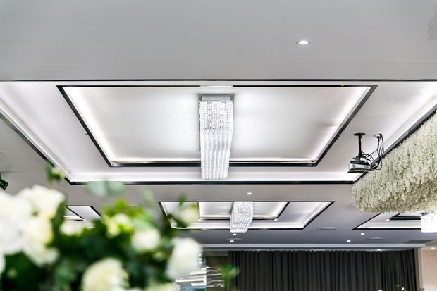 Upside-angled view of a refined interior ceiling featuring layered recessed rectangular ceiling coffers with concealed perimeter lighting, crystal pendant chandeliers, and a dark ceiling section with mounted equipment. A textured hanging greenery installation appears at the right, while blurred floral arrangements occupy the foreground. The space has a polished, event or hospitality-like atmosphere.
