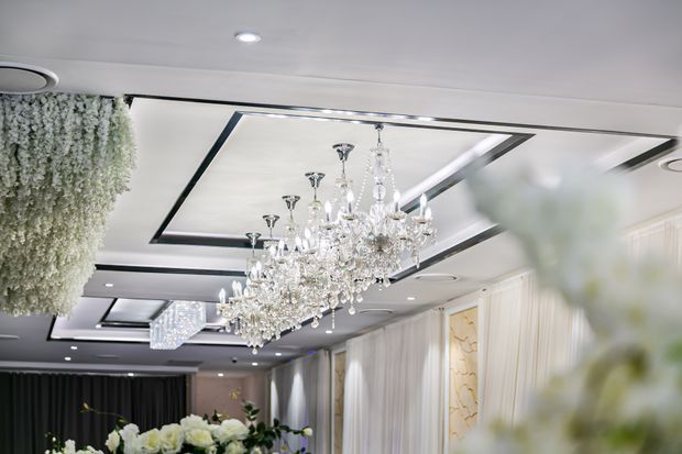 Elegant formal interior focused on a highly ornate crystal chandelier suspended from a coffered ceiling with dark trim. The space features recessed downlights, reflective ceiling panels, sheer drapery, decorative wall panels, and abundant white floral arrangements, creating a luxurious event or hospitality atmosphere.