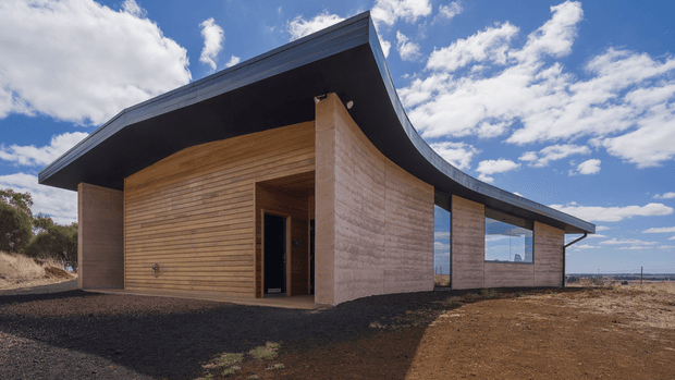 Exterior view of a distinctive single-storey contemporary building with curved, sweeping roof edges, a dark roof plane, warm horizontal timber cladding, curved textured earth-toned masonry walls, and extensive glazing. The structure sits on a sparse, rocky site beneath a bright blue sky.
