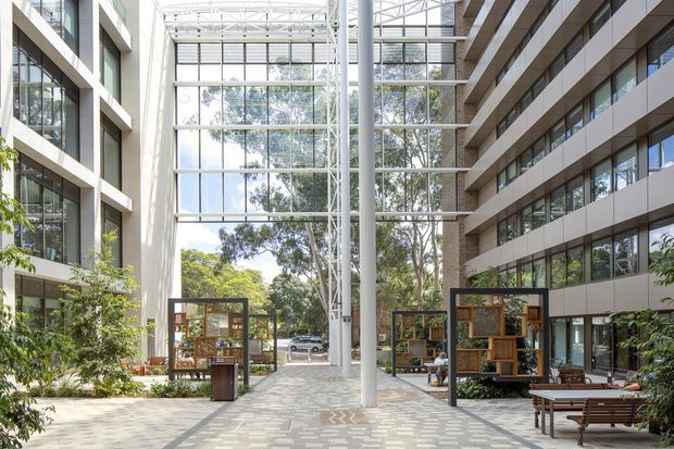 A bright contemporary commercial atrium or courtyard framed by multi-storey office buildings. The space features extensive floor-to-ceiling glazing, a large glazed roof and end wall, white exposed structural steel, pale patterned paving, landscaped planting, timber-and-glass display or seating structures, and outdoor benches. Mature trees and a street are visible beyond the glazed opening.
