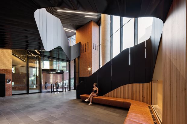 A dramatic contemporary commercial interior lobby with a spacious high-ceilinged entrance hall. The space features sweeping black sculptural ceiling and stair-like wall forms, extensive floor-to-ceiling glazing, warm timber wall cladding, orange-brown brickwork, dark metal detailing, and an integrated angular upholstered bench. Grey tiled flooring and linear architectural lighting complete the refined, high-contrast design.