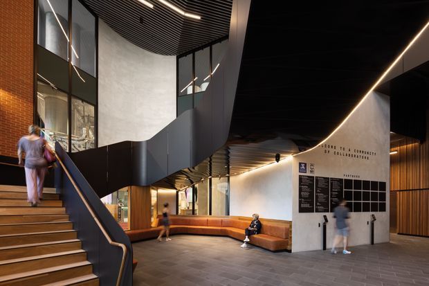 A dramatic contemporary interior lobby or public reception space with a broad staircase at left, black metal balustrades, exposed red brick, dark ribbed ceilings, linear integrated lighting, pale concrete or plaster walls, timber wall accents, and a long rust-coloured upholstered built-in bench. A large wayfinding or commemorative wall graphic is visible at right, with people using the space. The architecture features strong geometric forms, double-height glazing, and a polished institutional or cultural-building character.