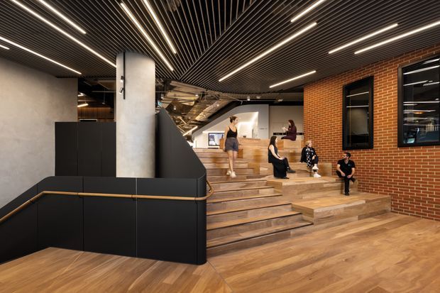 A contemporary multi-level commercial interior with broad timber steps forming informal seating and circulation. The space features a warm timber floor and stair platform, exposed red brick walls, dark acoustic slatted ceilings with linear integrated lighting, black metal joinery and balustrades, and a large cylindrical concrete column. Several people occupy the stepped area, with a reception or workspace visible in the background.