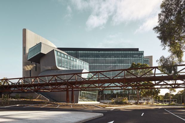 Exterior architectural photograph of a large contemporary institutional or commercial building with extensive curtain-wall glazing, a sculptural angular glazed volume, and a tall curved ribbed facade element. A rust-red steel pedestrian bridge crosses the foreground above a broad paved roadway and plaza.