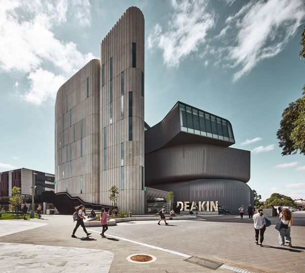 Exterior architectural photograph of a contemporary multi-storey institutional or university building. The complex features tall curved vertical volumes clad in ribbed light-grey metal, narrow vertical glazing, and a projecting dark sculptural wing with extensive clerestory-style windows. A broad paved pedestrian forecourt with people occupies the foreground, and the building signage reads “DEAKIN”.