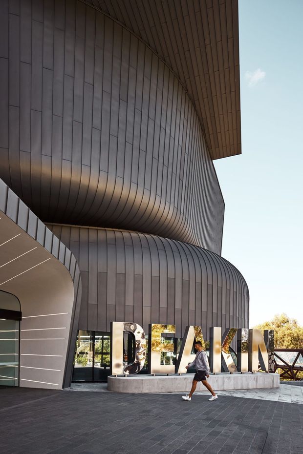 Exterior architectural photograph of a contemporary public building with dramatic sweeping, curved volumes clad in dark vertically segmented metal panels. Large freestanding entrance lettering sits on a concrete plinth, with a paved forecourt, glazed entry doors, and a projecting curved canopy. The composition emphasizes the sculptural facade and roofline against a clear sky.