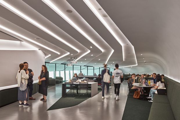 A large contemporary commercial interior, likely a café, lounge, or dining area, with a sculptural curved ceiling lined with layered recessed linear LED lighting. The space contains communal tables, booth seating, a long upholstered bench, people dining and socialising, and glazed partitions enclosing a meeting or work area. The palette is predominantly white, grey, charcoal, and black, with polished concrete-like flooring and a spacious open-plan layout.