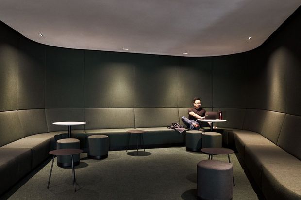 A dim, enclosed lounge or waiting area with curved built-in upholstered bench seating wrapping around dark acoustic-looking wall panels. Small round tables and cylindrical stools are arranged across a dark carpeted floor, with a person seated at a small work table near the back. Recessed ceiling lights create a subdued, intimate atmosphere.