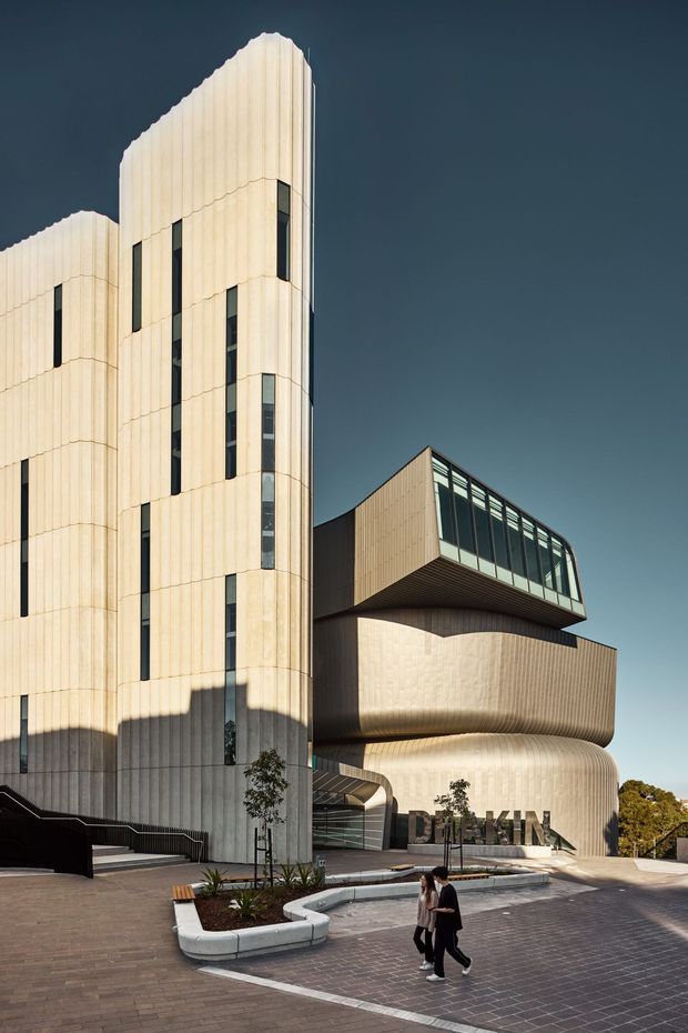 Exterior architectural photograph of a striking multi-storey contemporary building with pale ribbed or vertically grooved cladding, sculptural curved volumes, narrow vertical windows, and a projecting glazed upper-level form. The building fronts a paved pedestrian forecourt with raised planters and stairs; signage is visible on the rounded facade. Strong sunlight and deep shadows emphasize the geometric and organic forms.