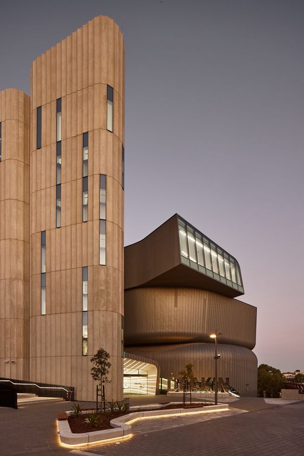 Dusk exterior view of a large contemporary multi-storey commercial building with sculptural curved volumes, tall timber-toned vertical cladding, narrow slit windows, and a projecting glazed upper-level volume. Illuminated pathways and a paved forecourt lead toward the recessed entrance, with landscaped planters at the base.