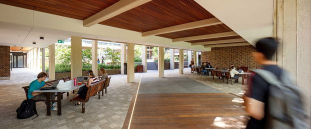 A spacious covered communal atrium or courtyard with open sides, brick feature walls, concrete-paver flooring, timber-lined ceiling panels and exposed white beams. Long tables with chairs provide informal study or dining areas, with people using laptops. The space connects to landscaped outdoor areas and appears to be part of a contemporary institutional or commercial building.