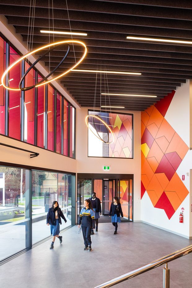 A spacious, brightly lit institutional entry lobby or atrium with full-height glazing along one side, polished concrete flooring, exposed dark ceiling beams, suspended linear strip lighting, and sculptural ring pendant lights. A vivid geometric mural in orange, pink, red, and yellow covers the rear wall, with students walking through the glazed entrance area and a stair handrail in the foreground.