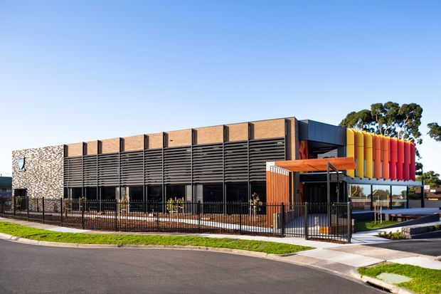 Street-level exterior photograph of a contemporary low-rise commercial building. The facade combines dark horizontal louvres, exposed warm brick panels, extensive glazing, a patterned masonry wall, and a vivid red-orange-yellow vertical fin feature beside the entrance. A fenced forecourt, pedestrian path, lawn, and paved driveway are visible, with clear blue sky and trees in the background.