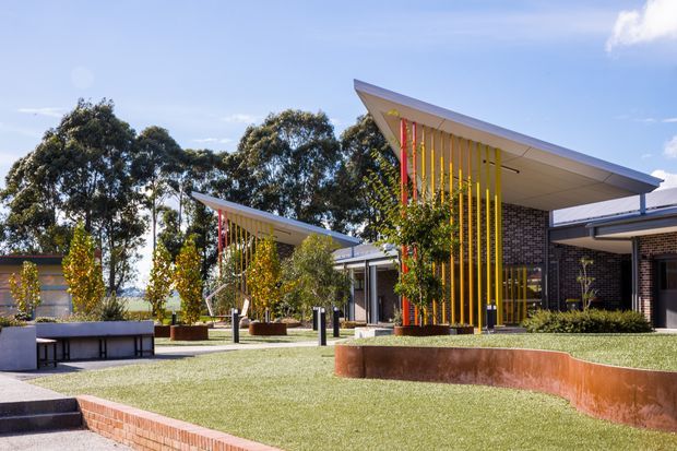 A contemporary low-rise building viewed from a landscaped courtyard. The architecture features broad asymmetrical sloping roof planes, dark brick walls, extensive glazing, and striking vertical yellow and red sun-shading fins at the entrances. The foreground includes manicured lawn, raised rust-coloured metal garden edging, brick paving, concrete seating, and mature trees under a bright blue sky.