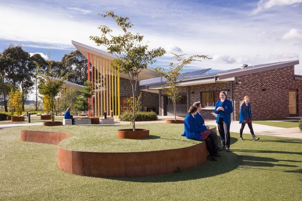 Exterior view of a contemporary brick institutional or educational building arranged around a landscaped courtyard. The complex features low-pitched roof forms, a projecting entrance canopy with vertical multicoloured fins, extensive glazing, paved paths, raised circular corten-coloured planter walls, and people gathered in the outdoor space.