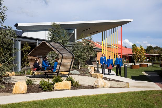 Exterior view of a contemporary institutional or commercial building with a broad low-pitched roof, extensive glazing, dark cladding, and a covered entrance marked by a vivid multicoloured vertical screen. Landscaped grounds include paved walkways, lawn, ornamental planting, large rocks, and a distinctive timber shelter with seating. People are present near the entrance and inside the shelter.
