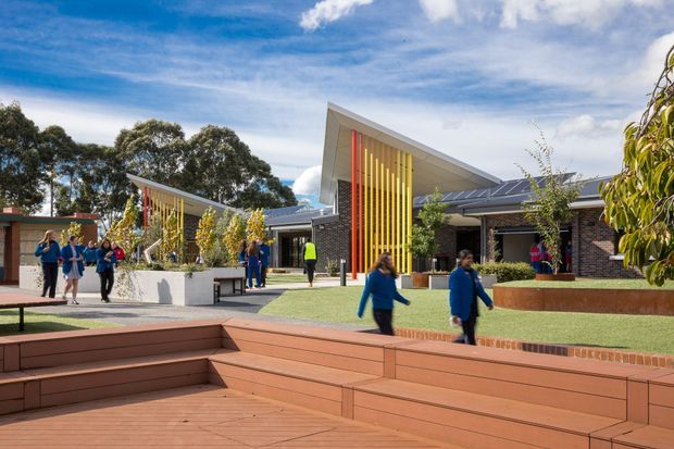 An outdoor courtyard and forecourt at a contemporary institutional or commercial complex. The scene features angular sloping roof forms, dark brick walls, prominent vertical yellow and orange sun-screen fins, landscaped planters, paved paths, and broad timber-toned stepped seating in the foreground. Several people occupy the courtyard, with mature trees and a bright sky in the background.