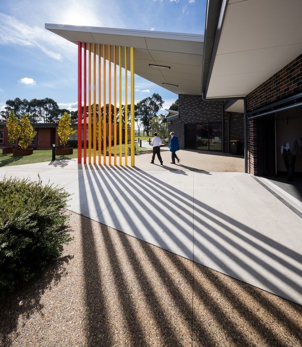 A bright contemporary architectural exterior showing a covered entry or courtyard walkway. A flat projecting roof is supported by a row of slender vertical red, orange and yellow fins that cast long graphic shadows across a pale concrete path and textured aggregate paving. Dark brown brick walls, white soffits, recessed linear lights and an open garage-like bay frame the right side, while landscaped planters and low buildings sit beyond. Two people provide scale.