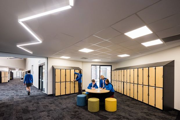 A bright contemporary school or institutional interior with banks of light timber lockers, dark patterned carpet, white acoustic ceiling tiles, recessed square lighting, and a small round table surrounded by colourful stools. People in blue clothing are present, and an adjacent corridor or open area is visible to the left.
