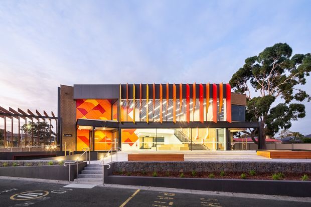 Exterior view of a contemporary two-storey commercial or institutional building with a bold geometric orange, red and yellow facade. Vertical orange and red fins screen the upper glazing, while large glazed openings reveal a bright interior. Brick side walls, dark metal framing, stepped concrete terraces, timber-toned benches and a covered pergola-like structure define the forecourt. The image is photographed from a paved parking area with mature trees in the background.