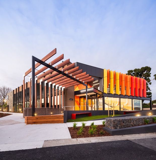 A contemporary low-rise commercial building with a bold, sculptural facade. The dark charcoal and brick exterior is framed by an open black steel canopy and a series of projecting timber-toned beams, while bright orange, yellow, and red vertical fins create a vivid feature wall around the glazed corner entrance. The site includes paved pedestrian areas, integrated planter beds, timber seating, gabion-style retaining elements, and low landscape planting. Photographed at dusk under a pale blue sky.